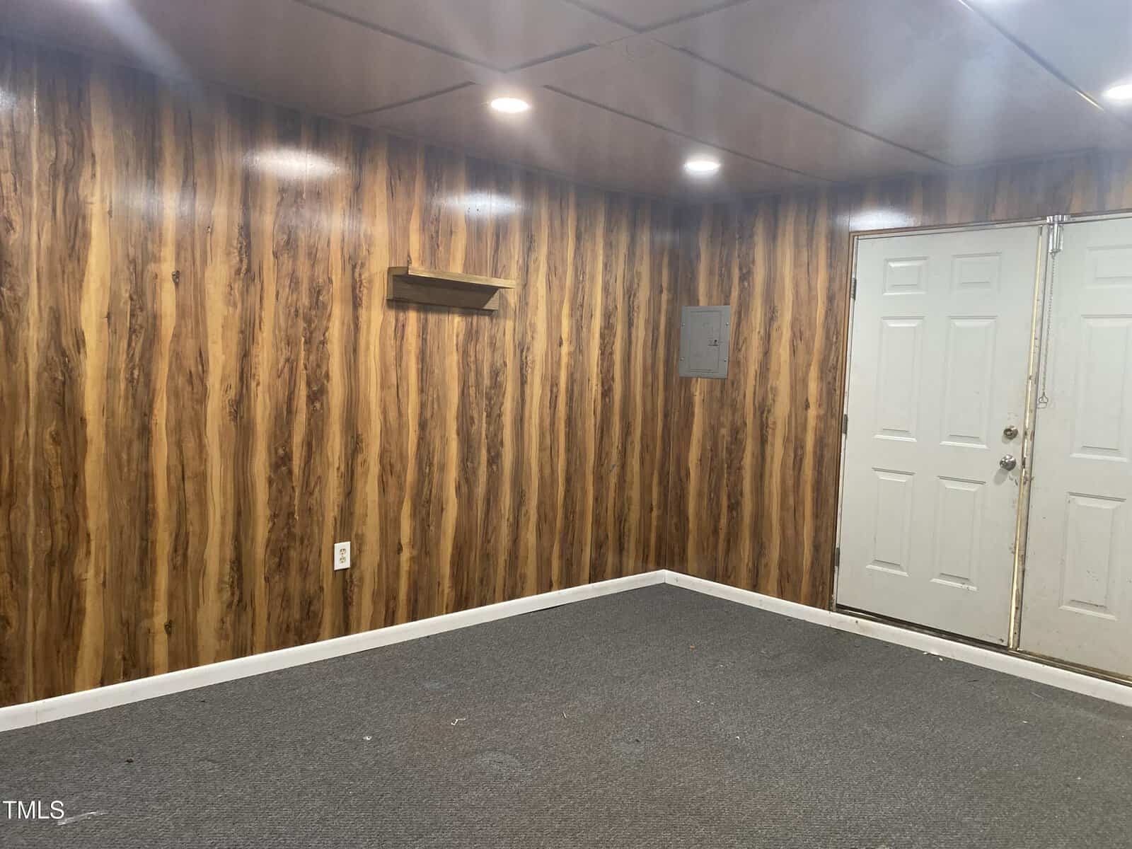 A small room with wood-paneled walls, gray carpet, a small wall shelf, an electrical panel, and double white doors. The ceiling has recessed lighting.
