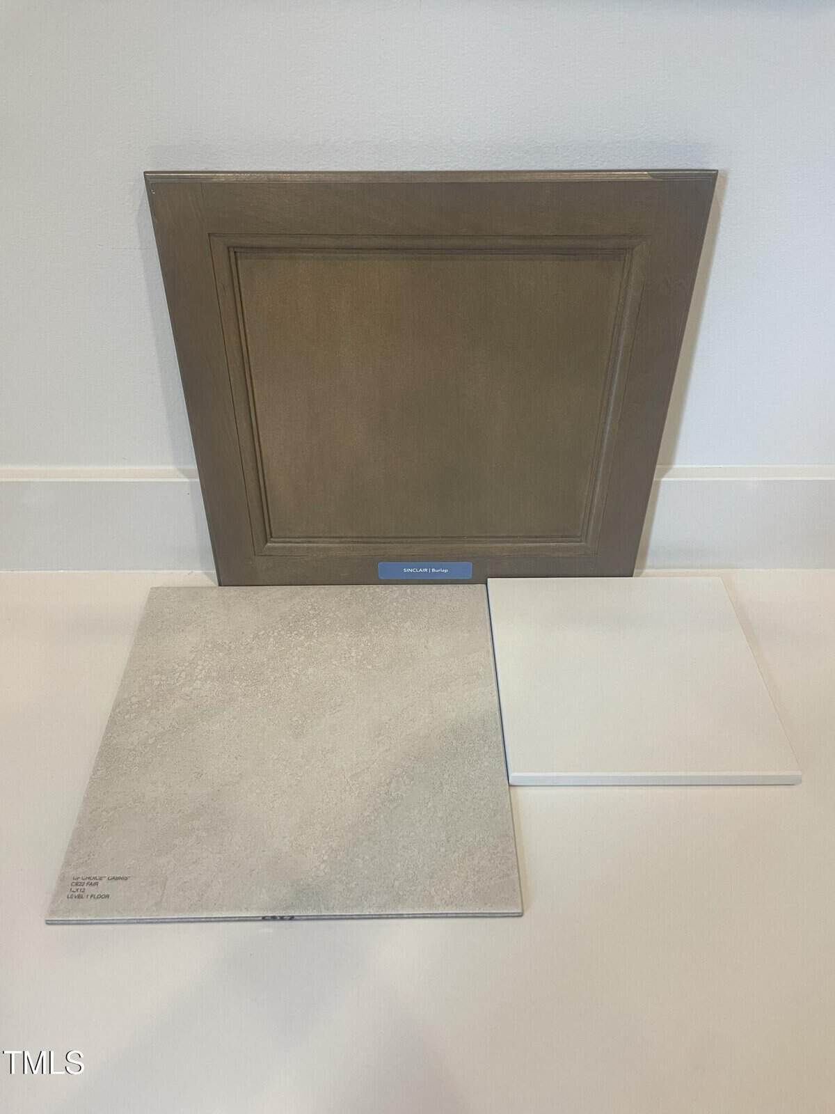 A wooden cabinet door sample stands against a wall. In front, there are two tile samples: one is light gray with subtle texture, and the other is solid white. All items rest on a white floor.