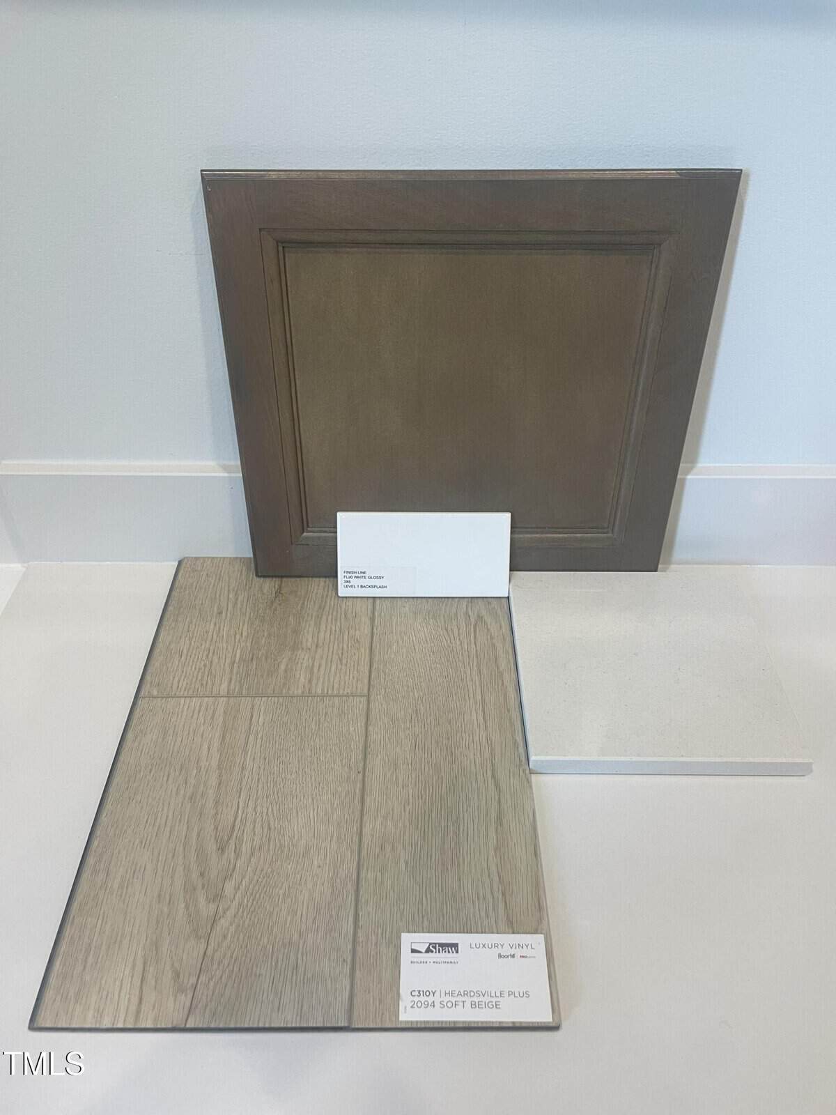 A dark wood cabinet door, a white paint swatch card, a light wood-look vinyl plank sample, and a square white tile are arranged upright and flat on a white surface against a white wall.