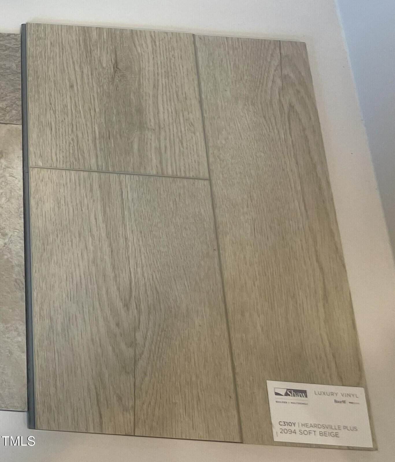 A sample board displays beige luxury vinyl flooring with a wood grain pattern. A label in the corner reads “Luxury Vinyl,” “C310Y Heardsville Plus,” and “20934 Soft Beige.” The flooring sample is rectangular.