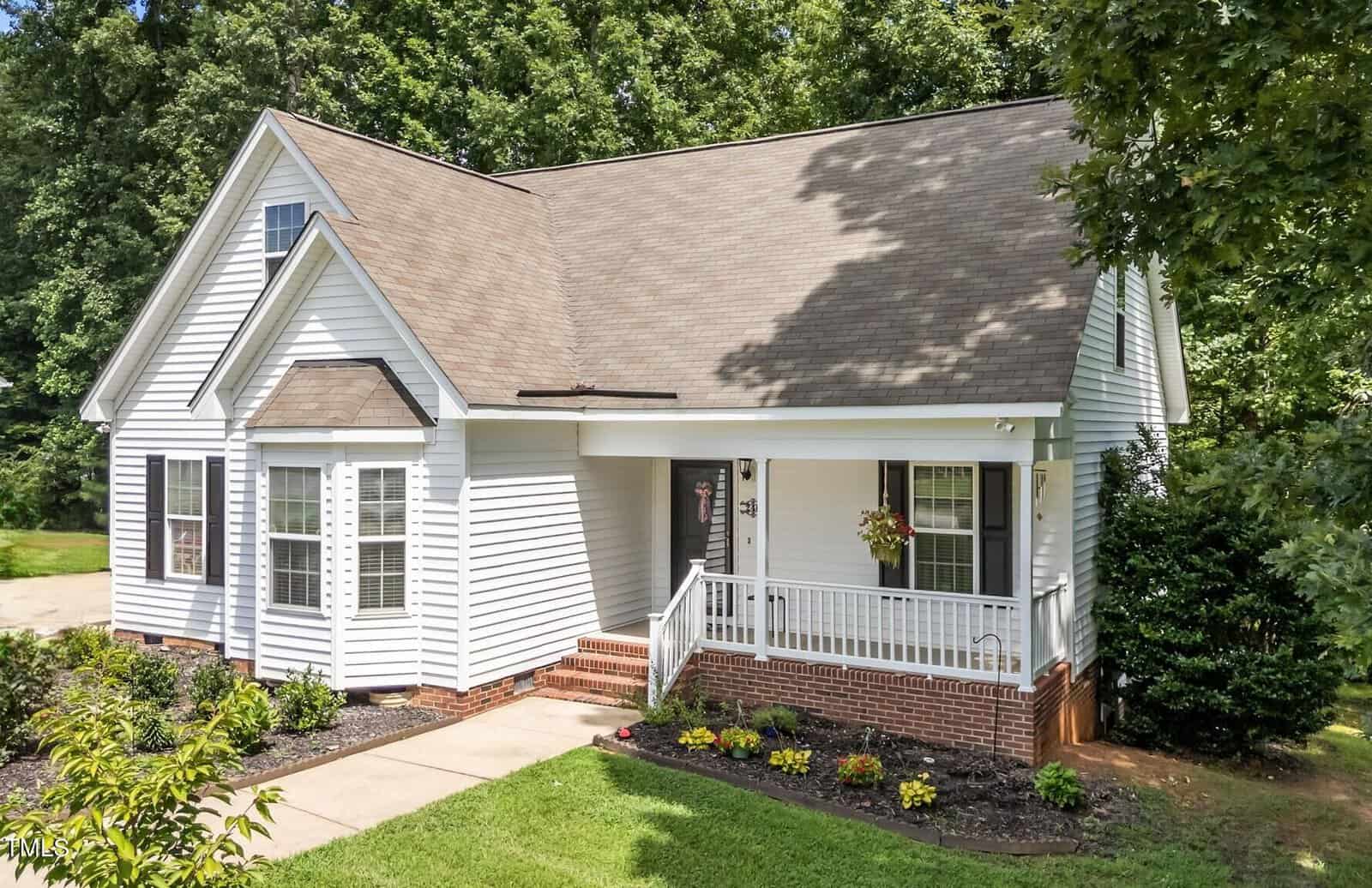 A white, single-story house with a gabled roof, front porch, and brick foundation sits surrounded by green grass, trees, and landscaped flower beds on a sunny day.