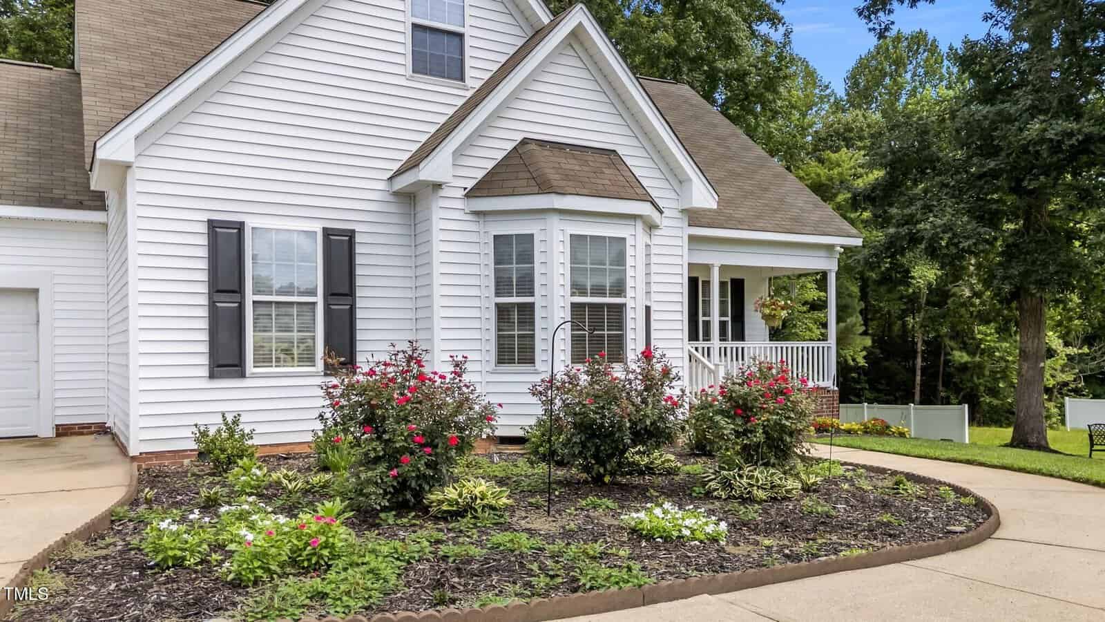 A white house with black shutters and a small front porch. There is a bay window and a landscaped garden with blooming flowers and shrubs bordered by a concrete driveway and green lawn. Trees are in the background.