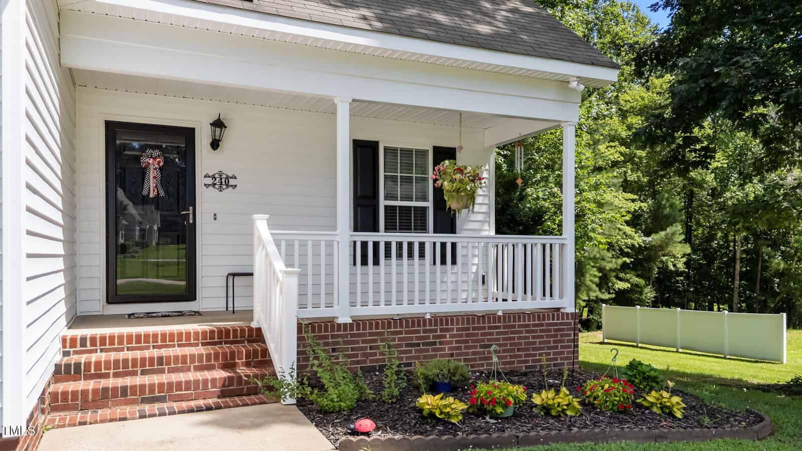 A white house with a front porch featuring white railings, a black door with a decorative wreath, brick steps, flower beds with colorful flowers, and a hanging planter, surrounded by green grass and trees.