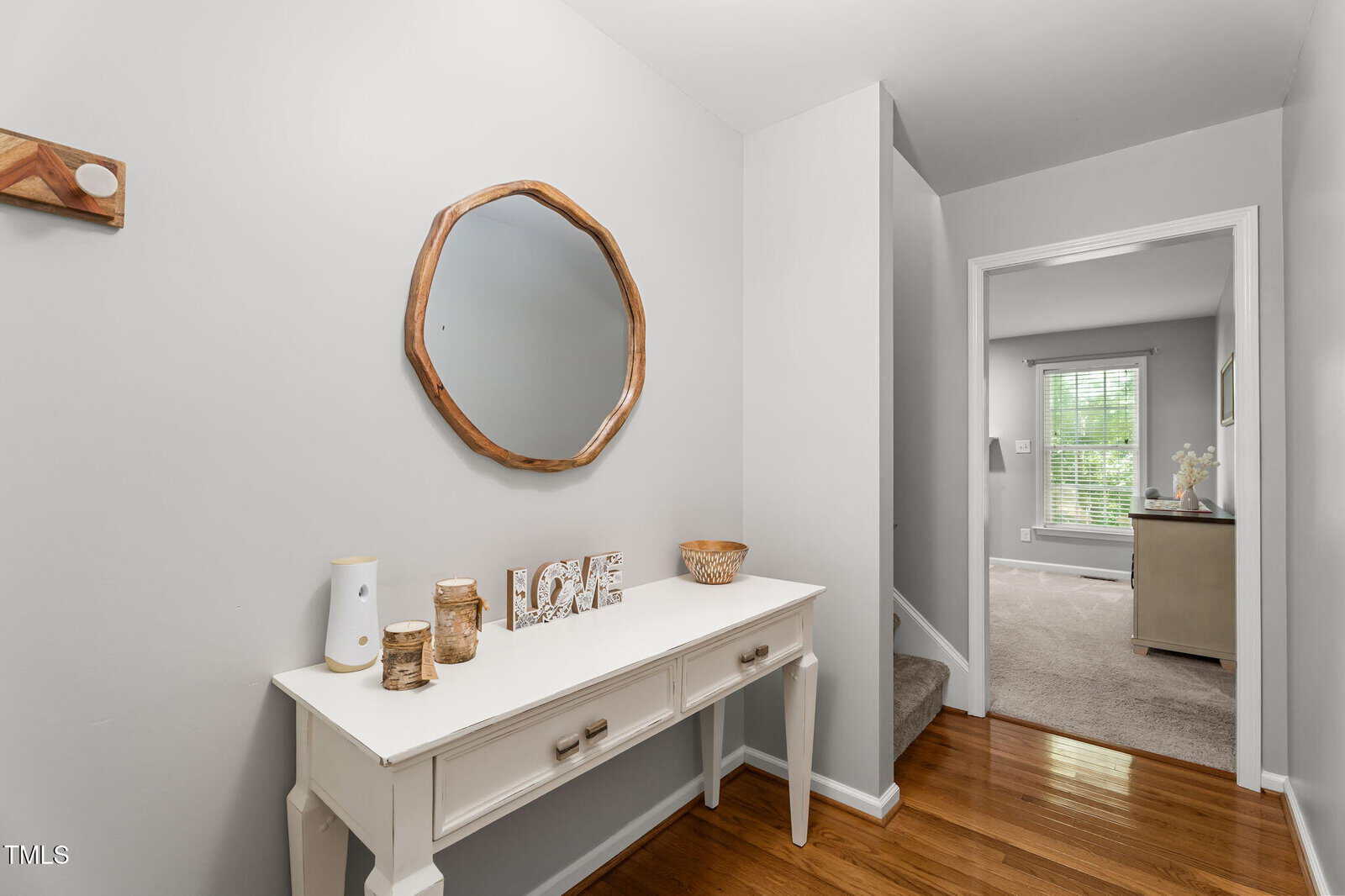 A hallway with light gray walls, a wooden floor, a white console table with decor, a round wall mirror, and a view into a carpeted room with a window.