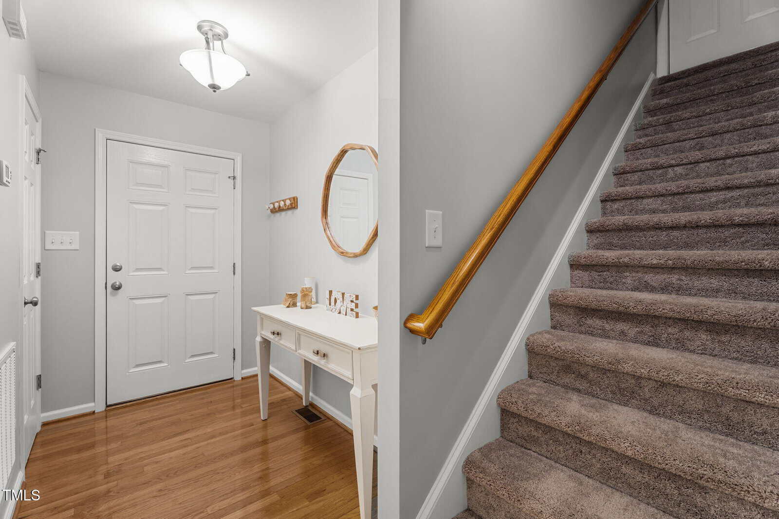 A foyer with a white door, a white console table with decor, a round mirror above it, and wood floors. Carpeted stairs with a wooden handrail are on the right side of the image.