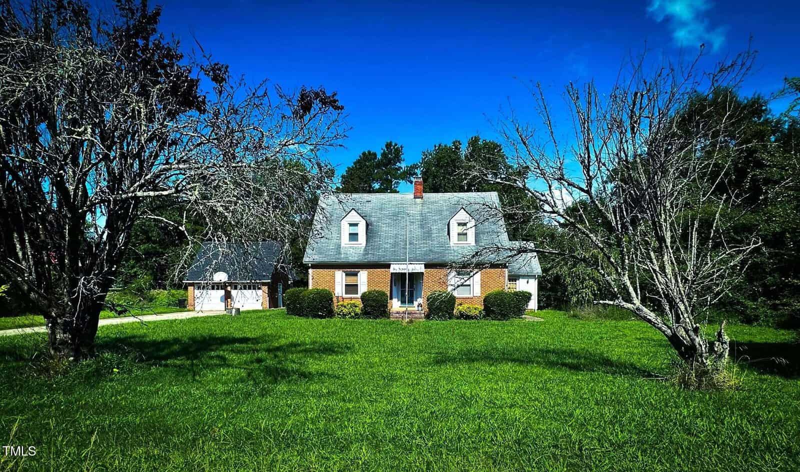 A two-story brick house with a steep roof and dormer windows sits on a large grassy lawn. Two leafless trees are in the foreground, with green bushes by the home's entrance and blue sky above.