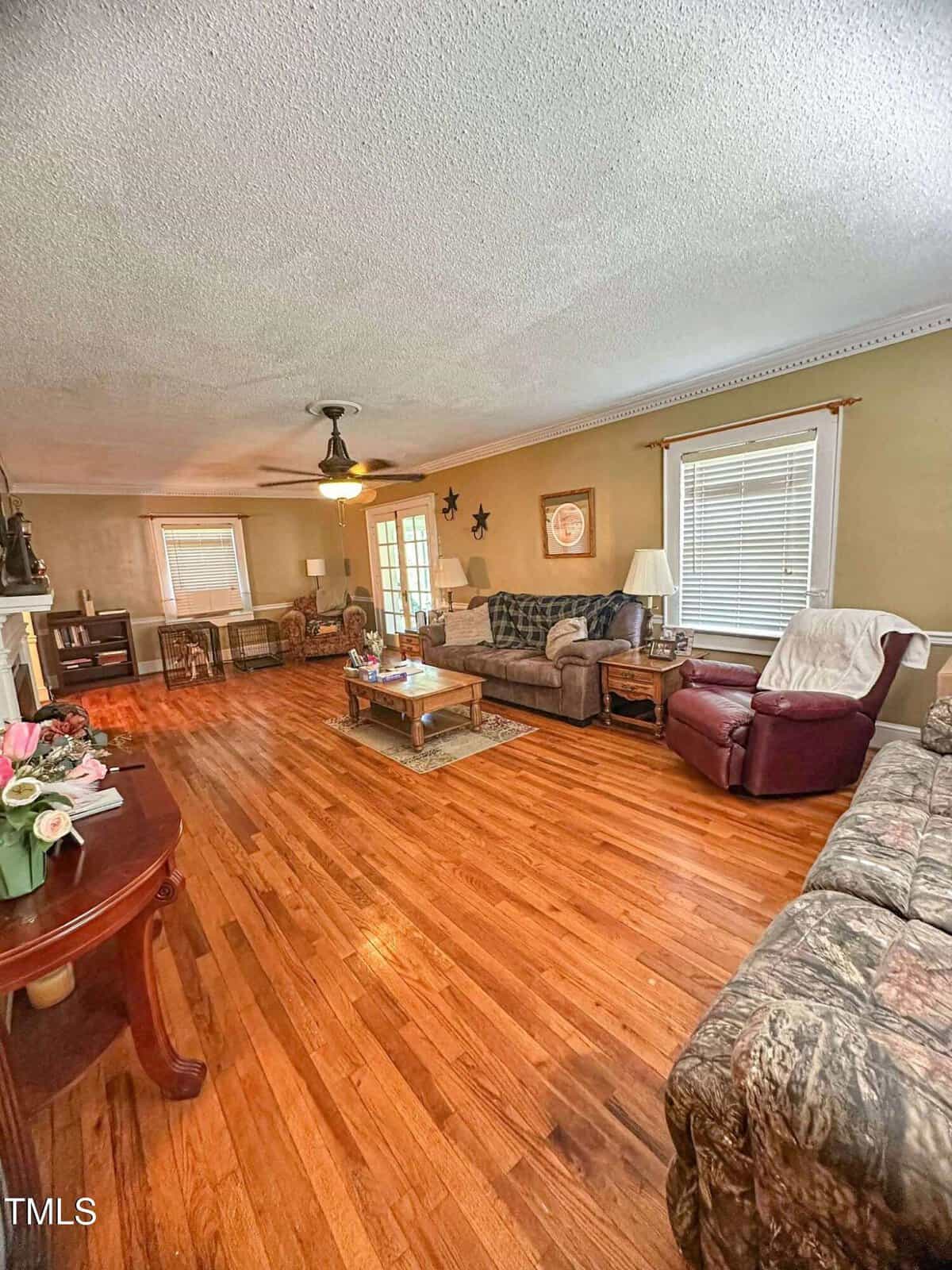 A spacious living room with hardwood floors, multiple sofas, a recliner, coffee table, standing lamps, and large windows with blinds. The room is decorated with wall art and has a ceiling fan with lights.