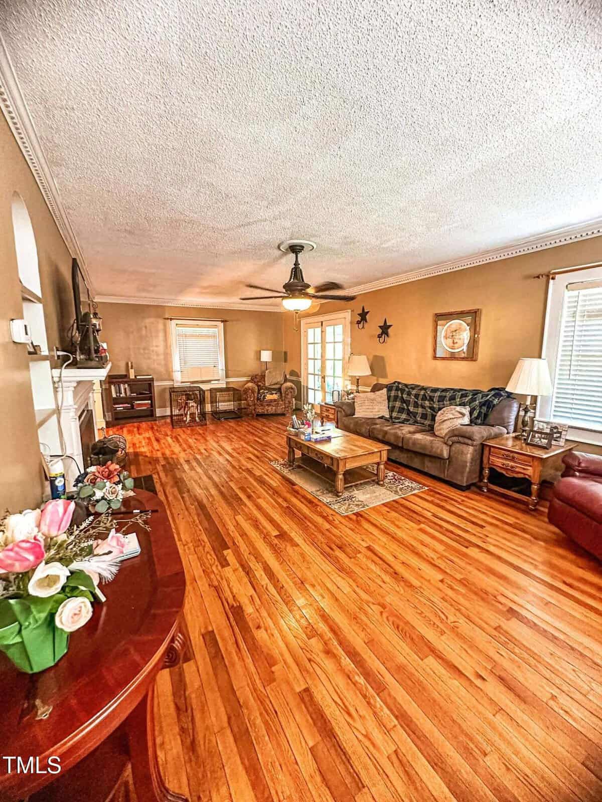 A spacious living room with hardwood floors, brown walls, a gray couch, coffee table, recliner, flowers on a side table, ceiling fan, fireplace, and large windows letting in natural light.