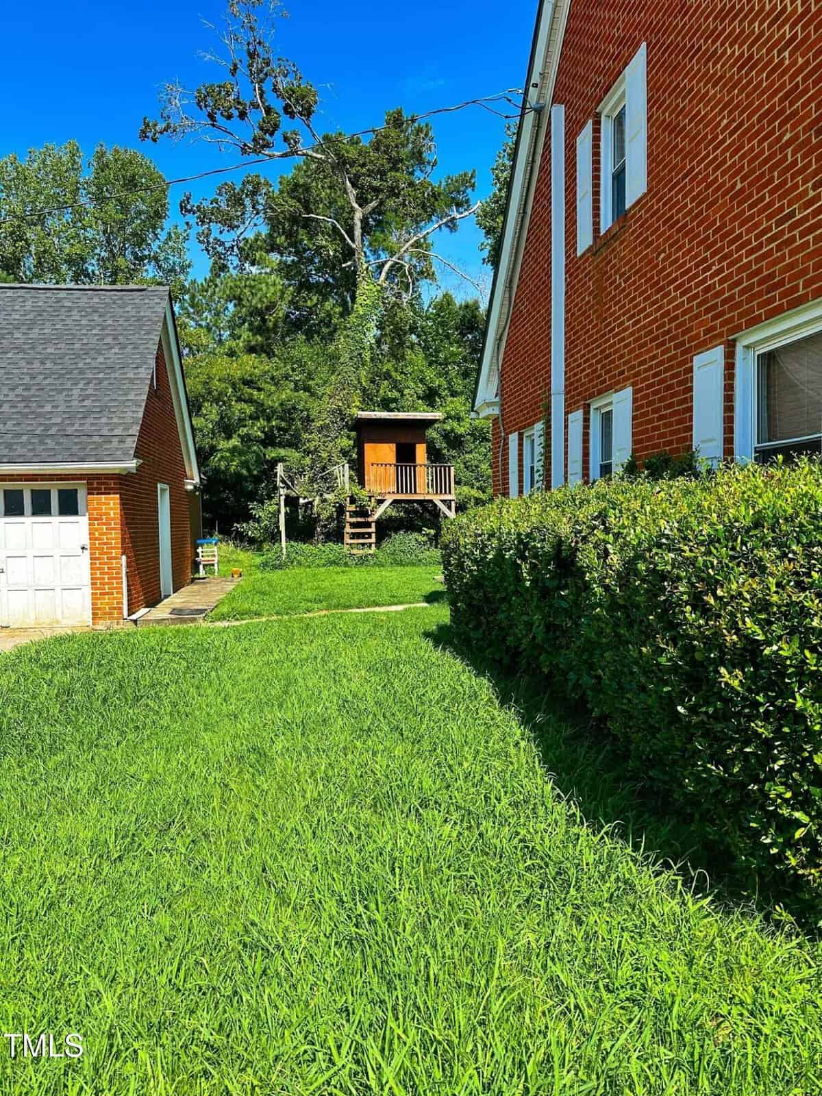 A green lawn runs between a brick house and a garage. In the background, a small wooden structure on stilts with stairs is visible, surrounded by trees and shrubs under a clear blue sky.