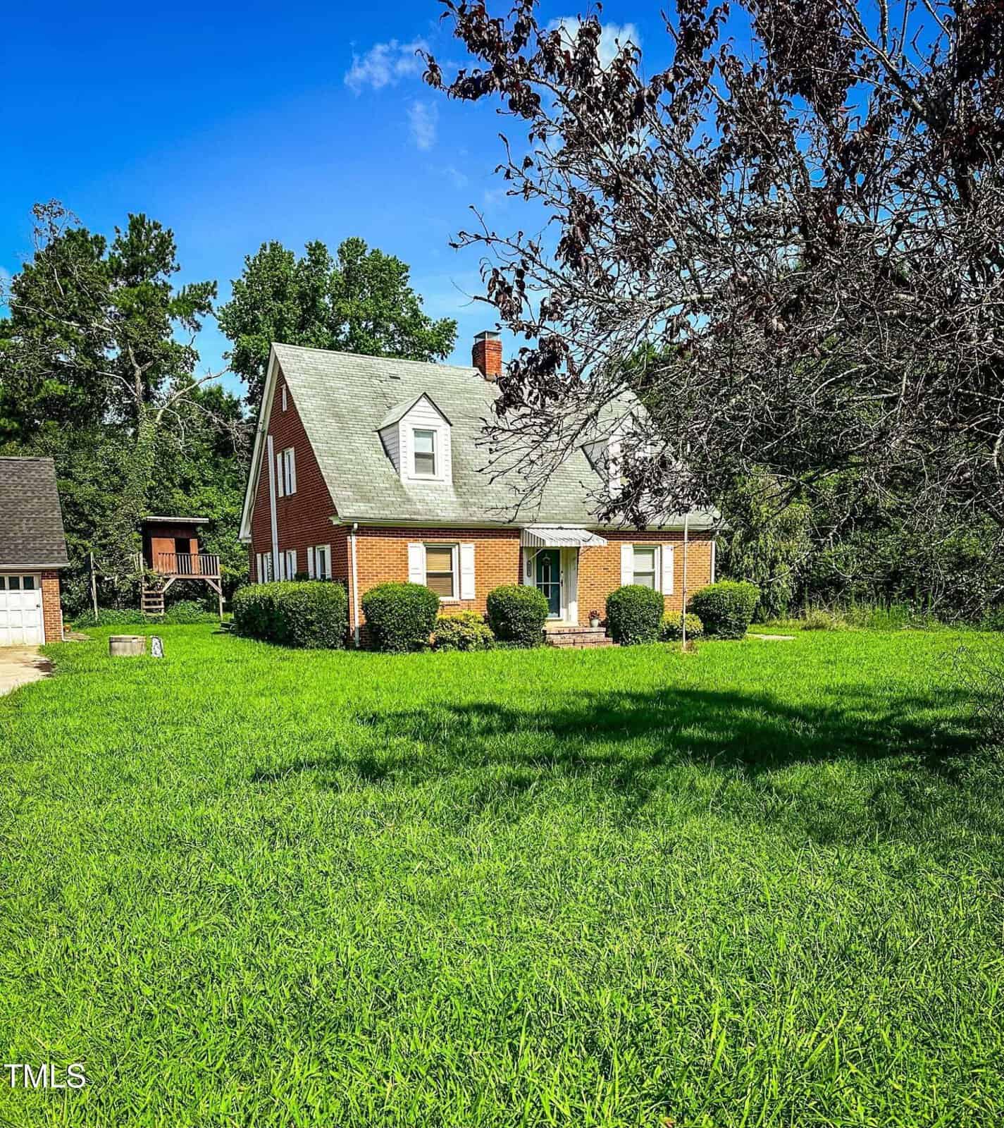 A red brick house with a green roof sits on a large, grassy lawn. The house has two dormer windows, a chimney, and shrubs lining the front. Trees are visible in the background under a clear blue sky.