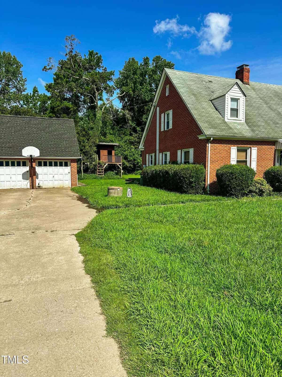 A red brick house with a gray roof sits beside a two-car garage with a basketball hoop. The driveway curves past a grassy yard, with trees and a wooden playset in the background under a blue sky.