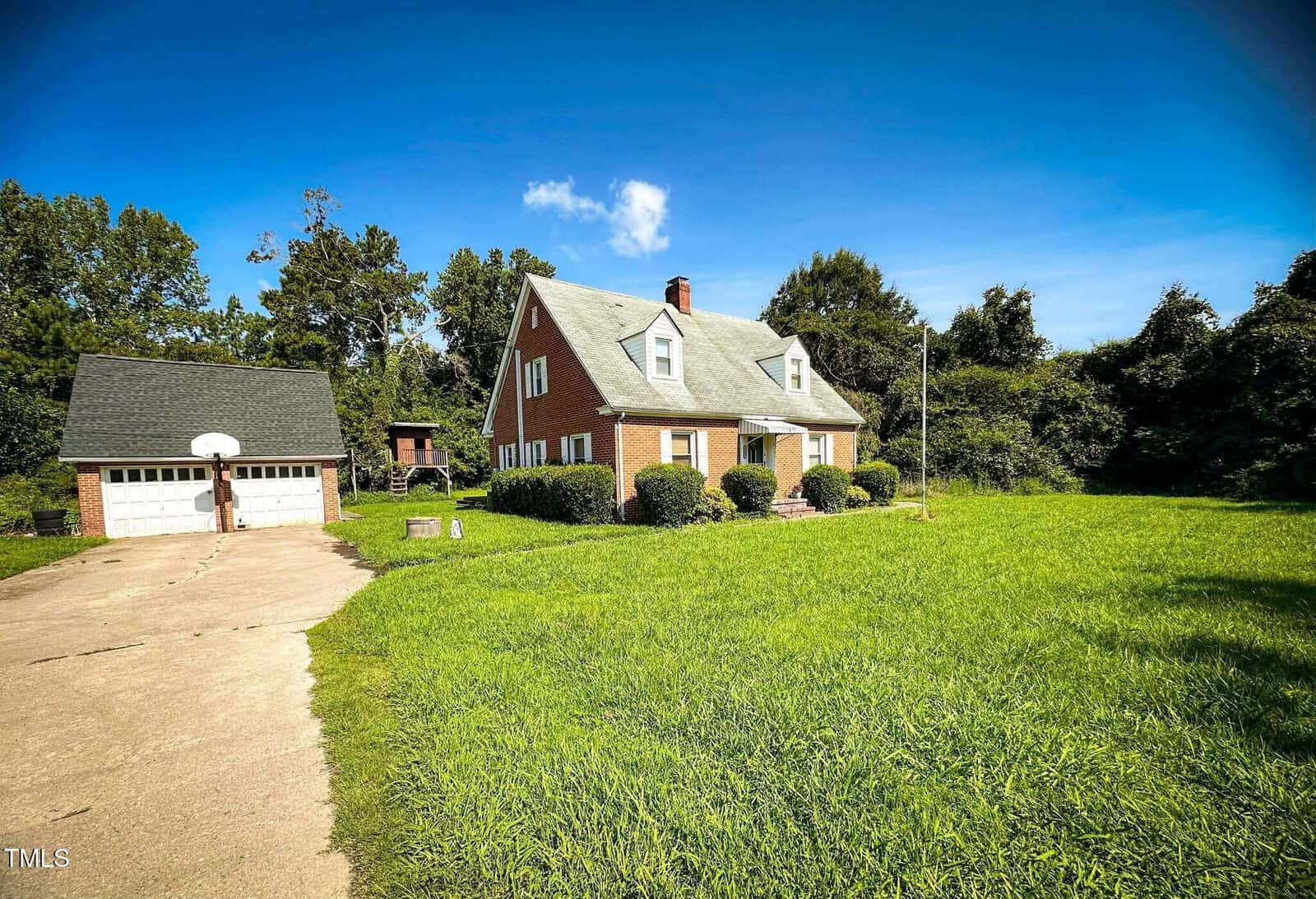 A two-story brick house with white trim and a steep gray roof sits beside a detached two-car garage at 8683 US Highway 15 in Bullock. The property features a long driveway, green lawn, and trees in the background under a blue sky.