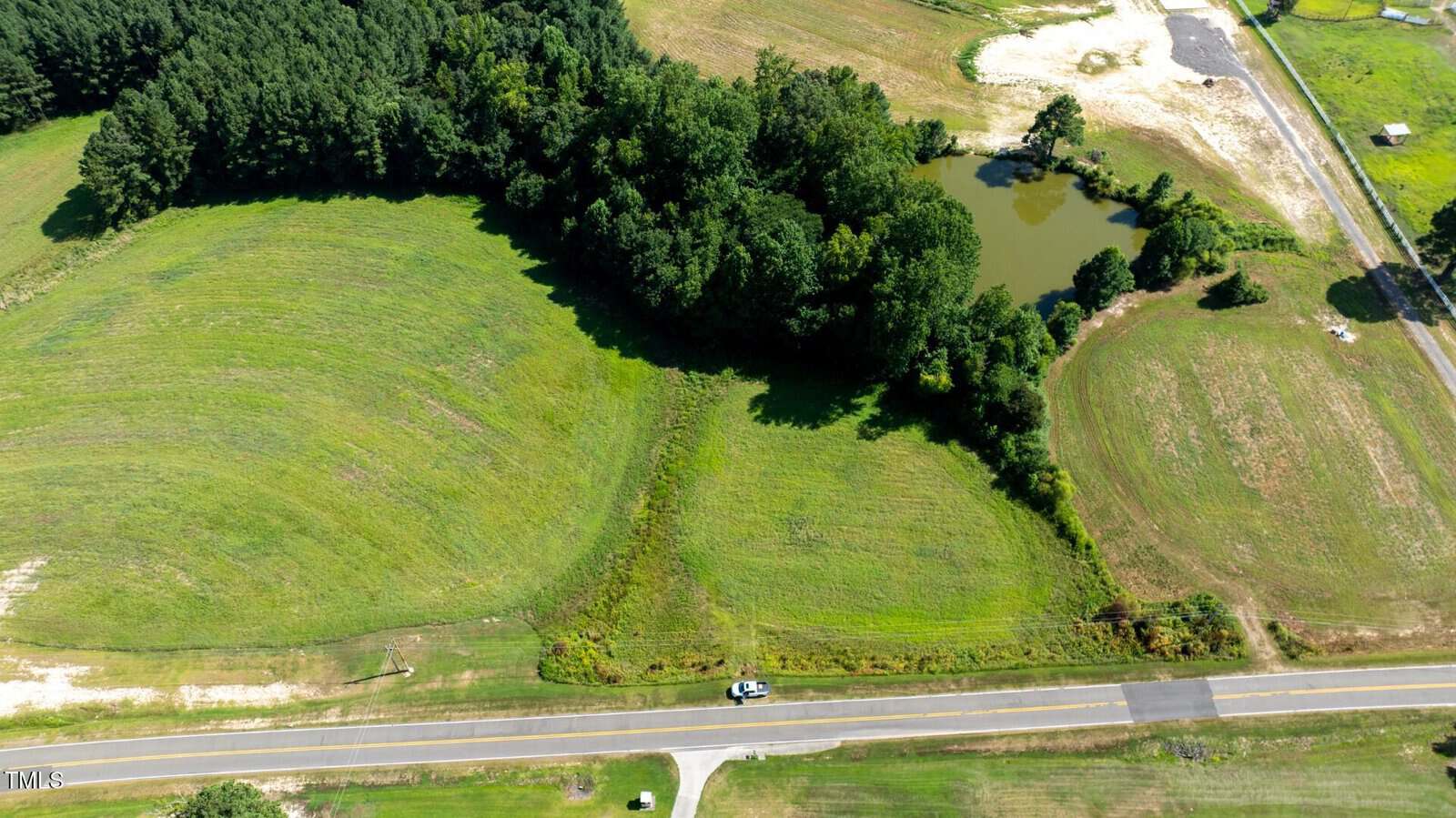 Aerial view of a landscape with a two-lane road, grassy fields, a small pond surrounded by trees, and a single vehicle parked on the roadside. A wooded area borders part of the fields.