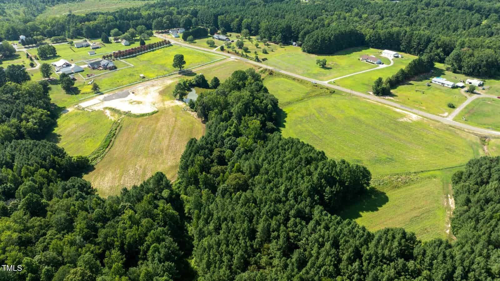 Aerial view of a rural area with open grassy fields, clusters of trees, a few houses, and intersecting roads under clear daylight. Some forested sections border the fields.