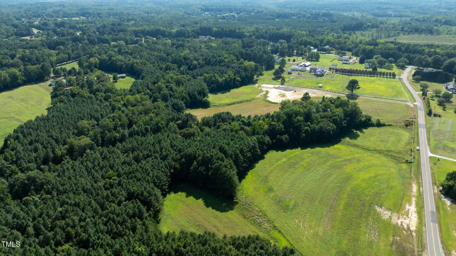 Aerial view of a rural landscape with dense forested areas, open grassy fields, scattered houses, and a road running along the right side of the image under a clear sky.