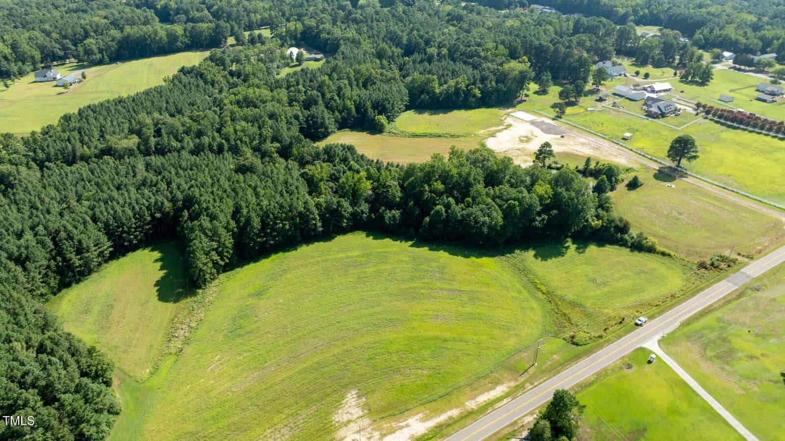 Aerial view of a rural area with green fields, patches of forest, scattered houses, and a road running along the edge of the scene on a sunny day.