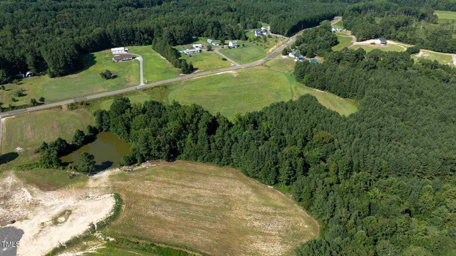 Aerial view of a rural area with open fields, a small pond, scattered houses, a road, and dense clusters of green trees surrounding the properties.