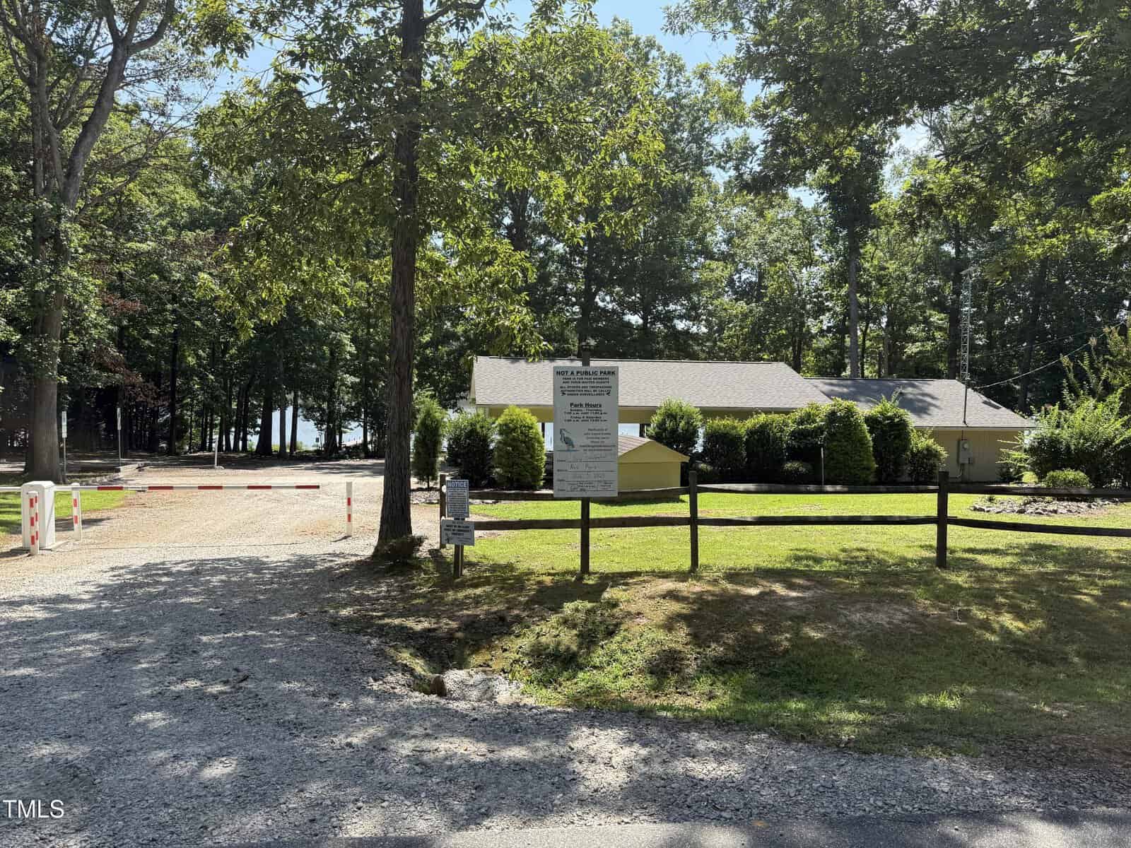 A gravel driveway leads to a small building surrounded by trees at 0 Summer Place, Macon. A wooden fence and a sign are in front, and a lake is visible in the background. Sunlight casts dappled shadows on the ground.