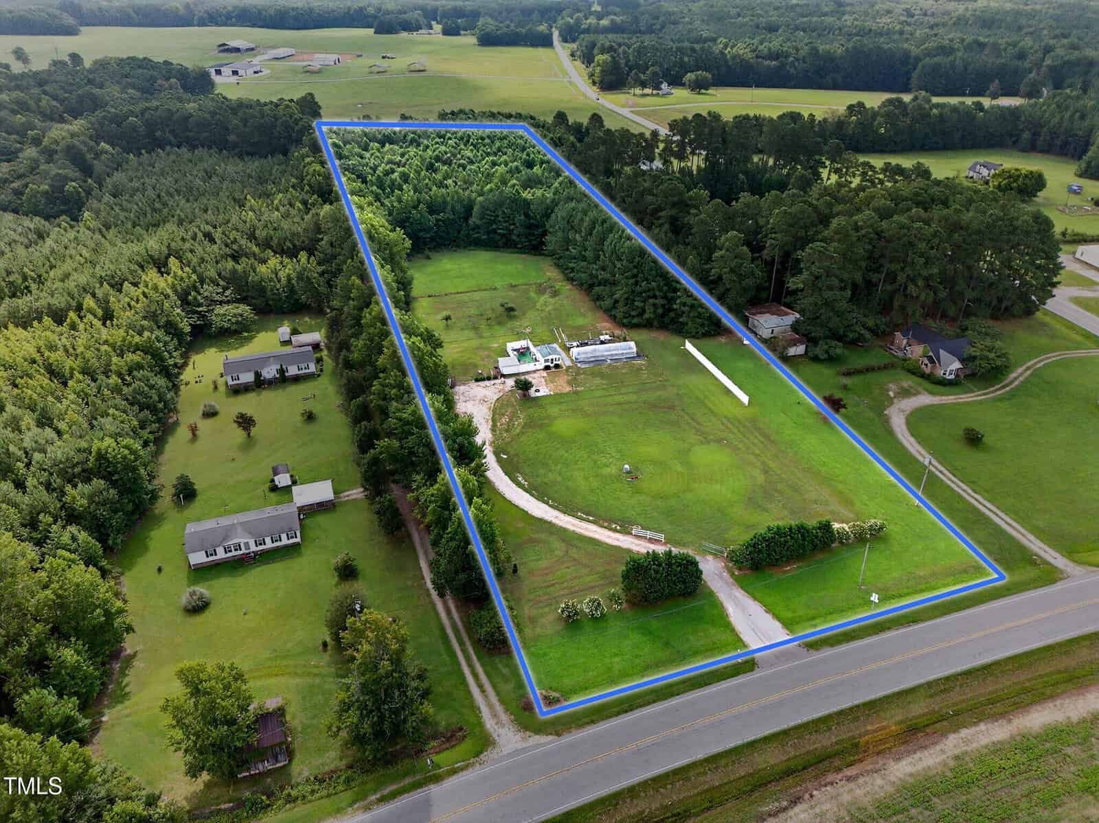 Aerial view of a rectangular, fenced rural property with a house, outbuildings, a curved driveway, trees, and open green space, bordered by a road and surrounded by other houses and forested areas.