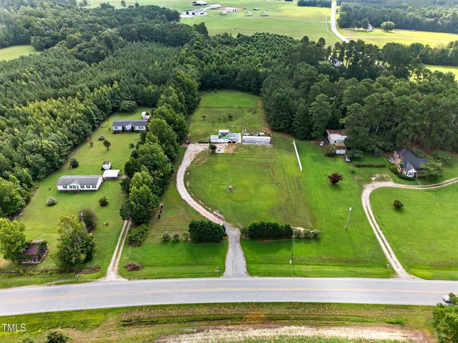 Aerial view of a rural property with several houses, green lawns, trees, a curved driveway, and a road in the foreground. Fields and wooded areas surround the homes. The landscape appears open and well-maintained.
