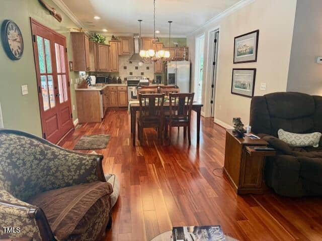 A cozy living and dining area with wood flooring, brown furniture, a dining table with four chairs, an armchair, a patterned chair, and a view into a kitchen with wooden cabinets and white appliances.
