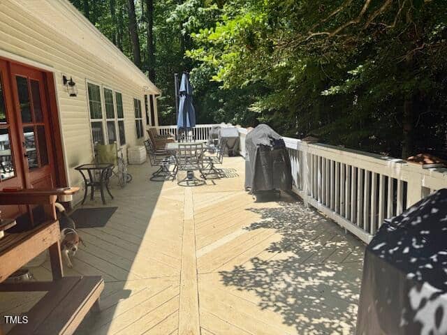 A spacious wooden deck with outdoor furniture, two grills, and an umbrella, attached to a house. Sunlight and shadows from trees cover parts of the deck. A small dog walks near the house.
