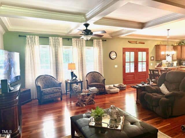 A living room with hardwood floors, two upholstered chairs, and a recliner facing a TV. A glass coffee table sits in the center, and French doors lead to the kitchen area. Light streams in from multiple windows with curtains.