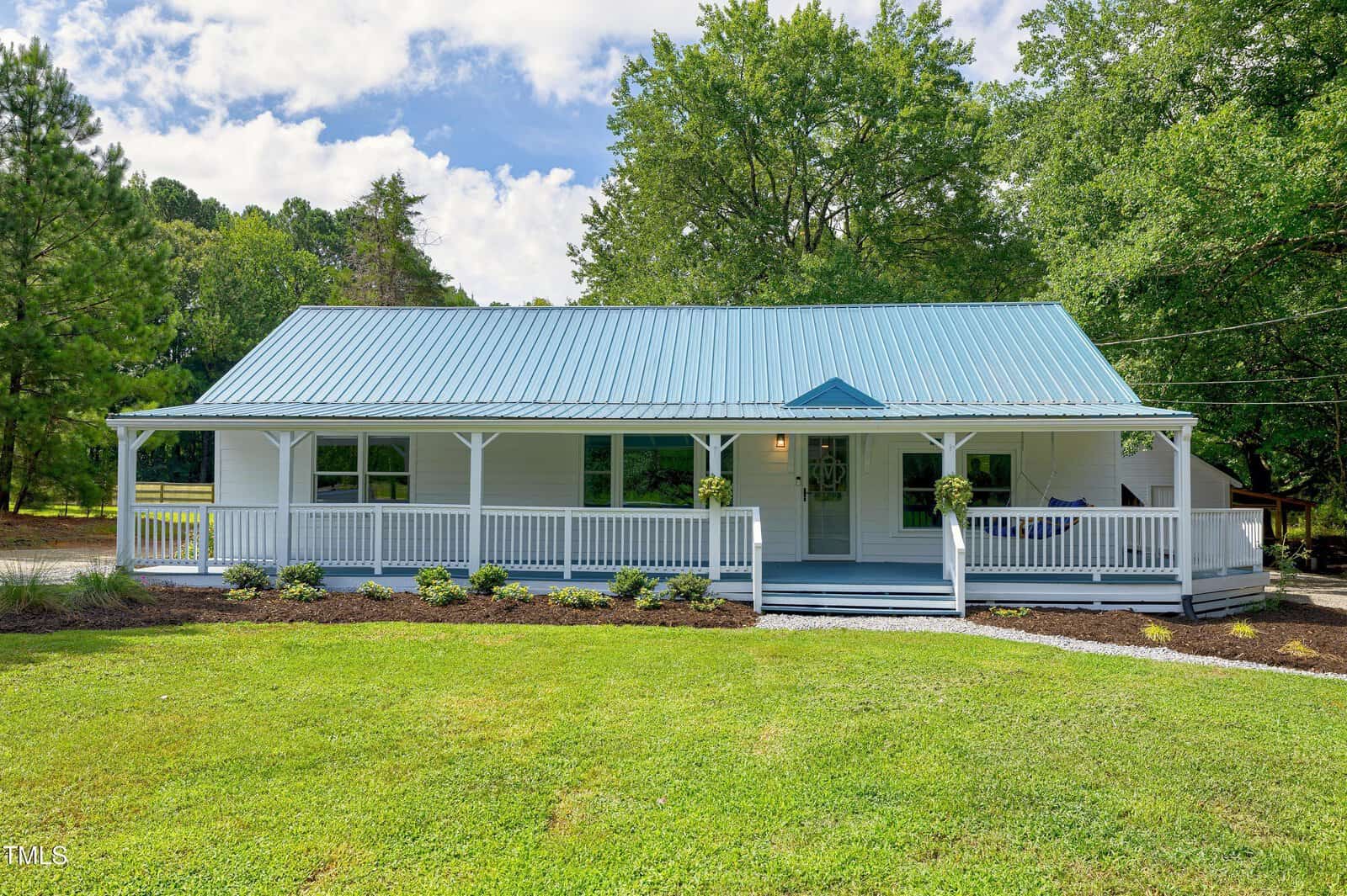 A single-story house with a blue metal roof, white siding, and a wide covered front porch. The porch is bordered by white railings, and the house is surrounded by green grass and trees.