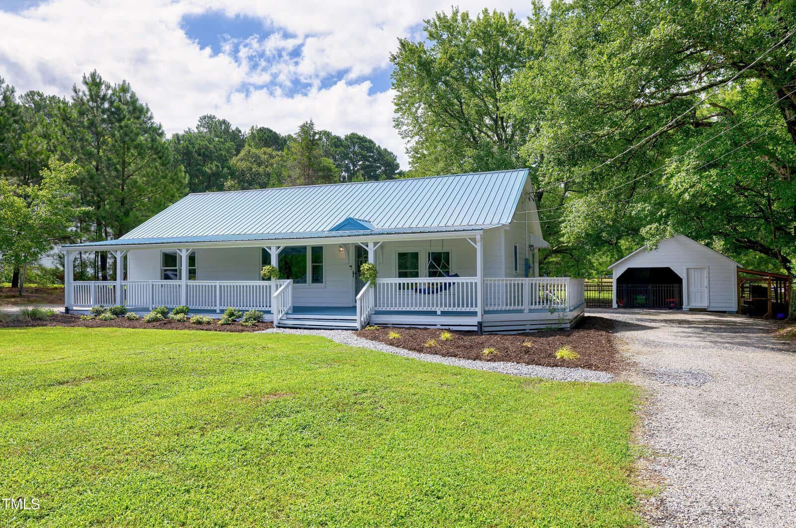 A charming white single-story house at 2542 Nc-56, Creedmoor, features a spacious front porch, metal roof, and lush greenery. A gravel driveway leads to a detached garage, with trees and lawn under a partly cloudy sky.