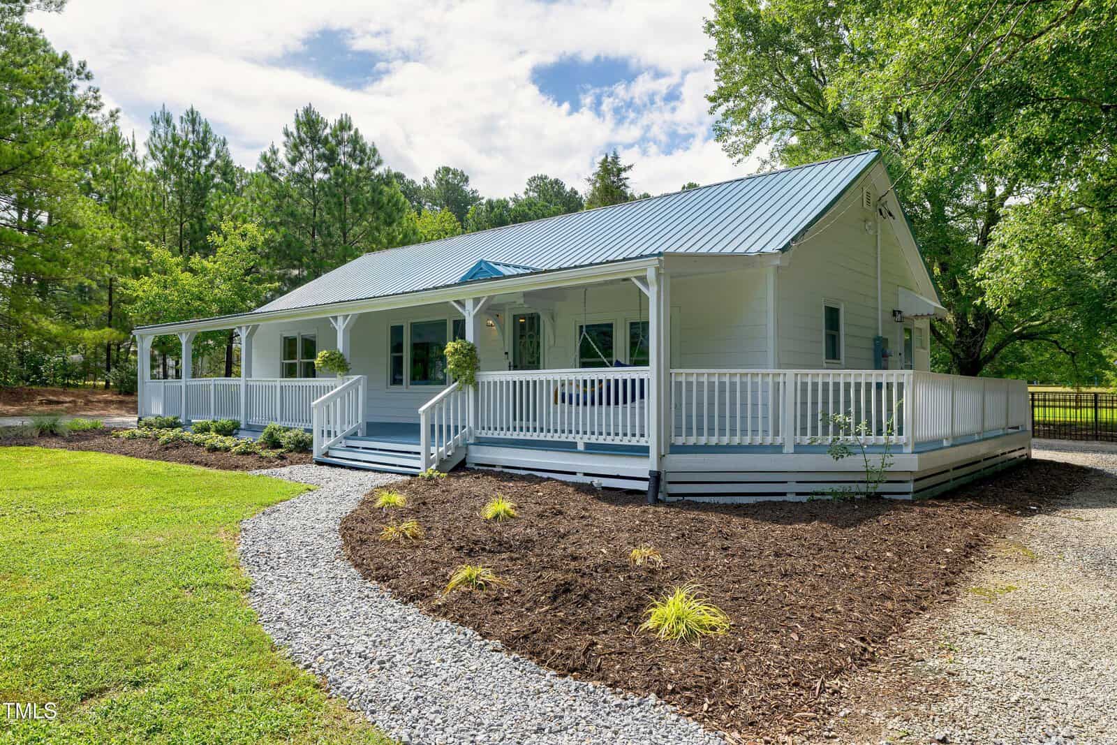 A white single-story house with a blue metal roof and wraparound porch sits amid green grass, landscaped mulch beds, and tall trees under a partly cloudy sky. A gravel path leads to the front steps.