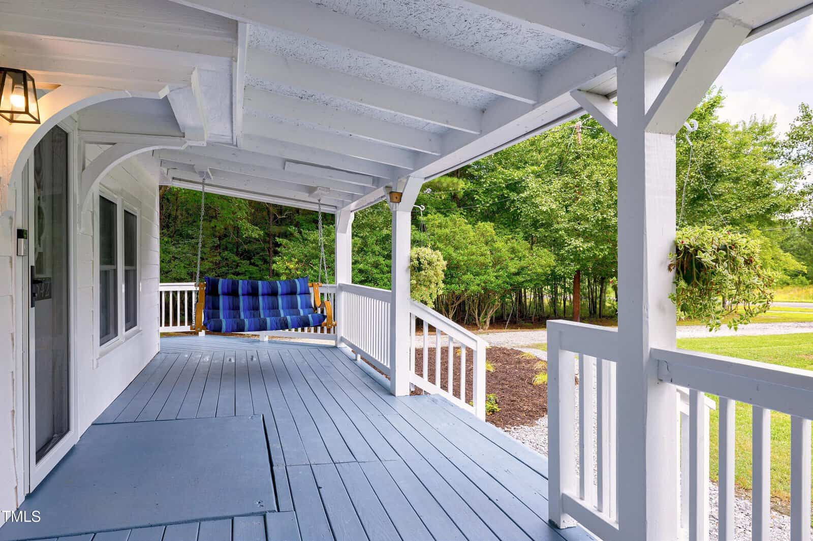 A covered porch with light blue wooden flooring, white railings, and a blue cushioned porch swing. Trees and greenery are visible in the background.