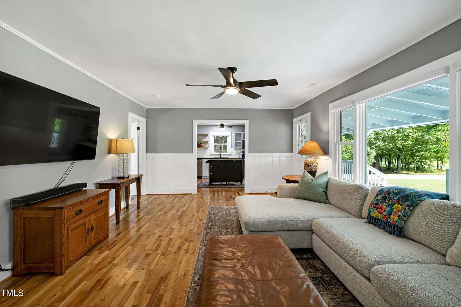A living room with hardwood floors, a sectional sofa, a large wall-mounted TV, wooden furniture, two lamps, a ceiling fan, and large windows overlooking a porch and green yard.
