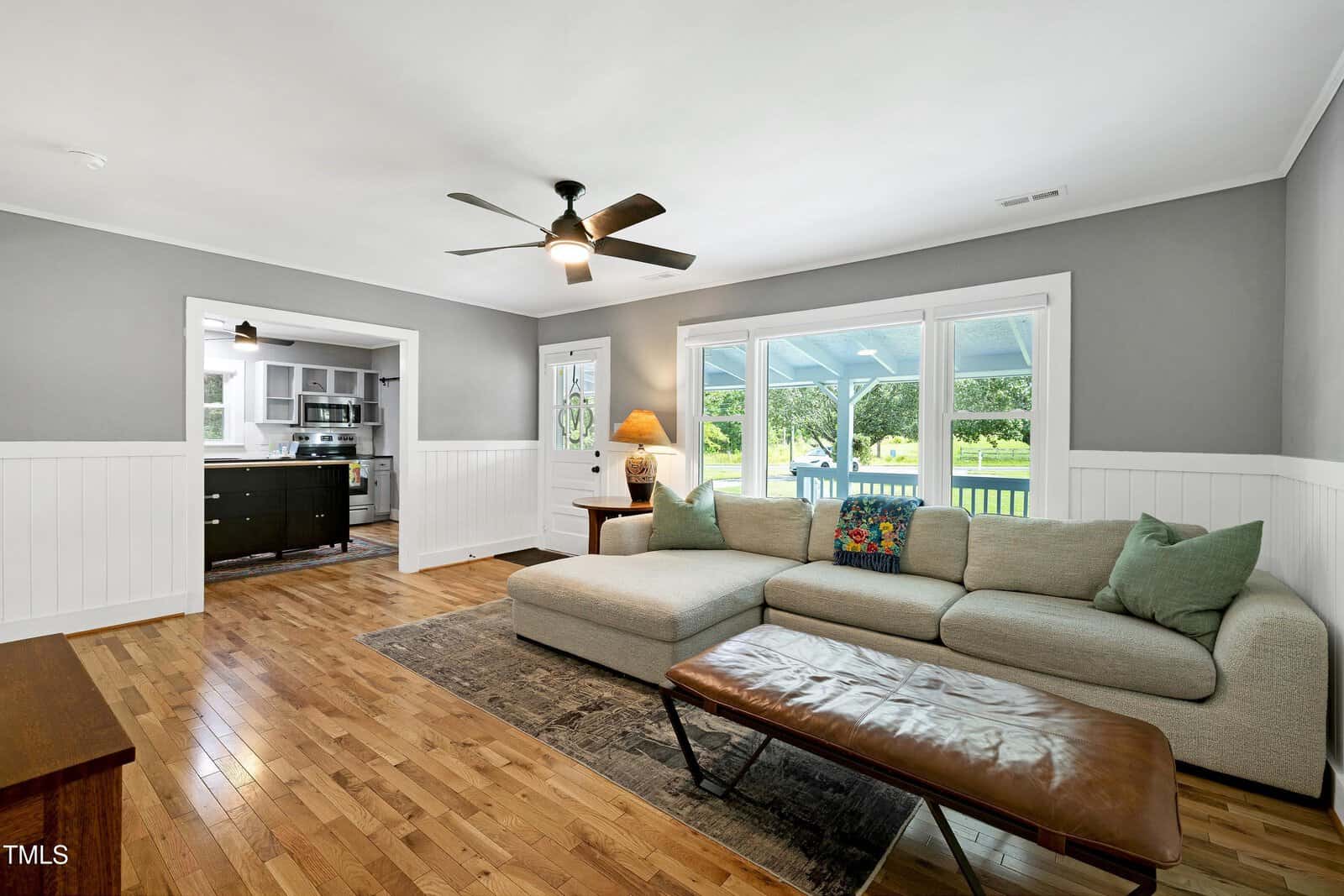 A living room with light gray walls, wood flooring, a beige sectional sofa, a leather bench, and large windows. The space opens to a kitchen area in the background, and a ceiling fan is mounted above.