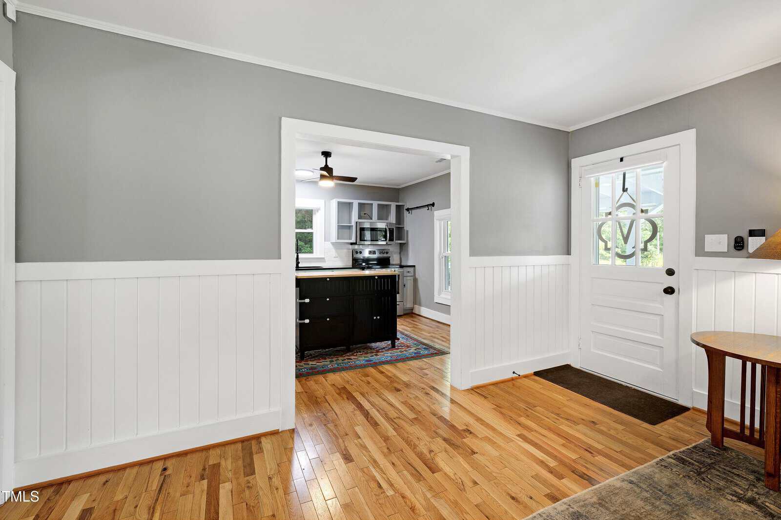 A view of a bright entryway with wood floors, white paneled walls, and a door with glass panels, leading to a kitchen with gray walls, an island, and stainless steel appliances.