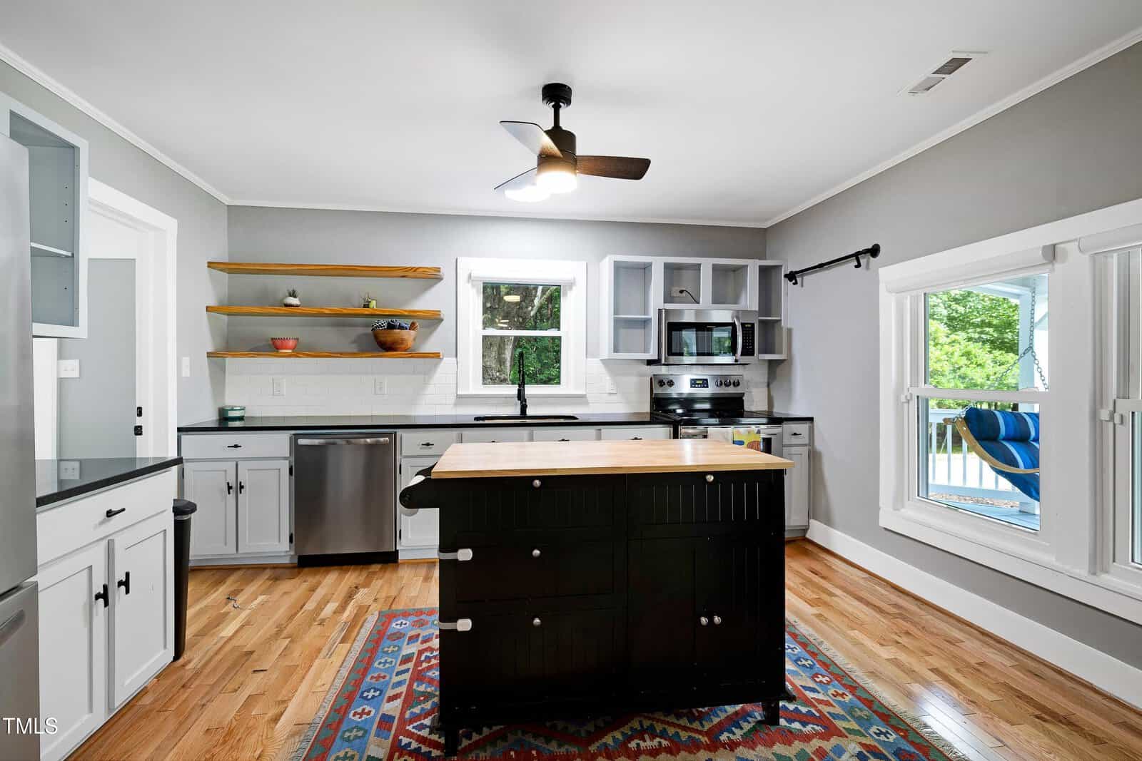 Modern kitchen with gray cabinets, stainless steel appliances, open wooden shelves, a central black island with drawers, hardwood floors, a colorful rug, and large windows letting in natural light.