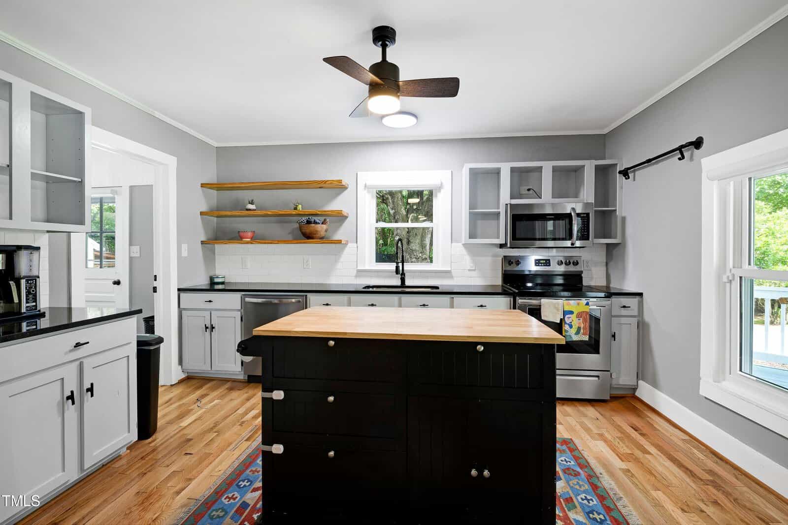 Modern kitchen with gray cabinets, black island, stainless steel appliances, wooden shelves, and light wood flooring. A window above the sink and another on the right bring in natural light. Ceiling fan and recessed lights are visible.