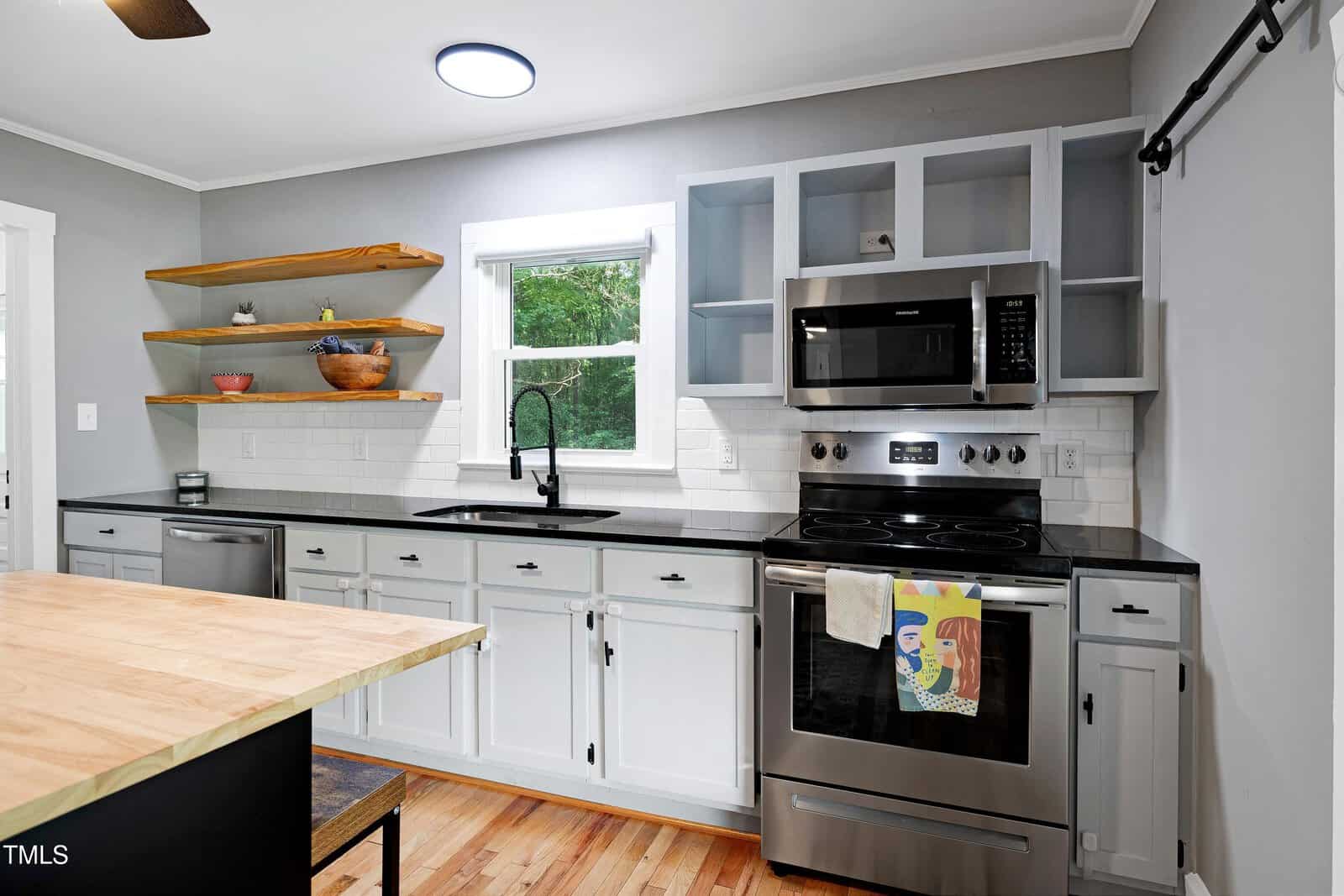 Modern kitchen with gray cabinets, stainless steel appliances, open wood shelves, a black faucet, white subway tile backsplash, and a window above the sink. A towel with an abstract design hangs on the oven handle.