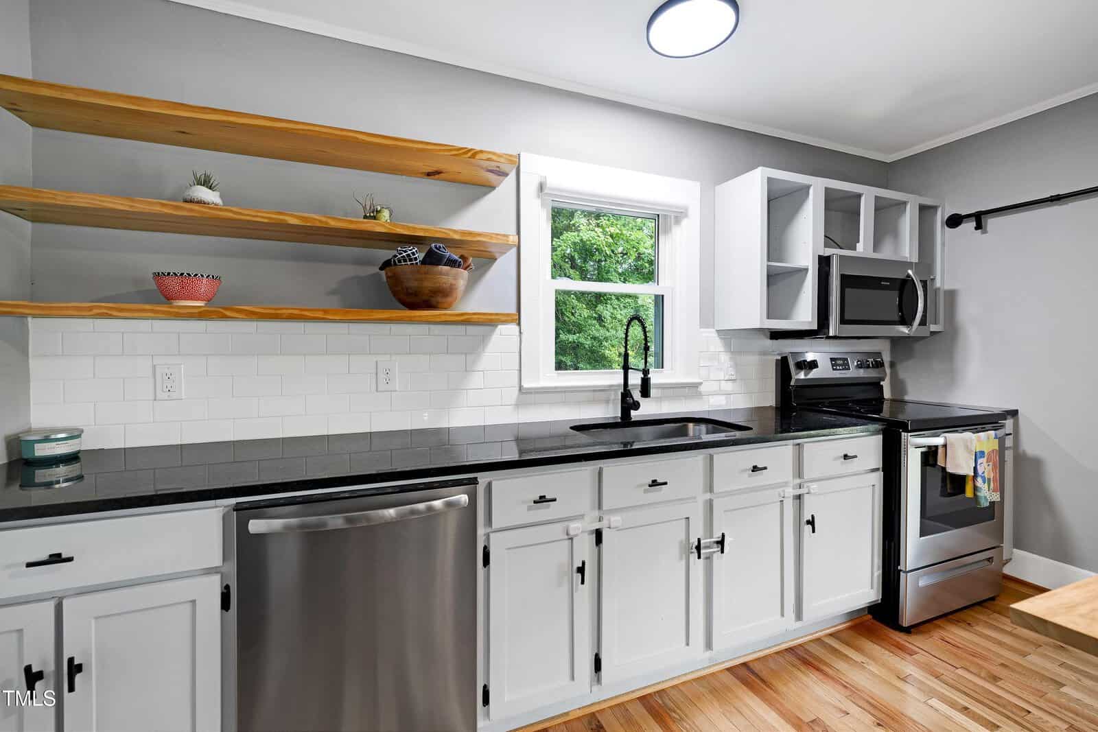 Modern kitchen with white cabinets, black countertop, stainless steel appliances, open wooden shelves with decor, a white subway tile backsplash, and a window above the sink showing green trees outside.