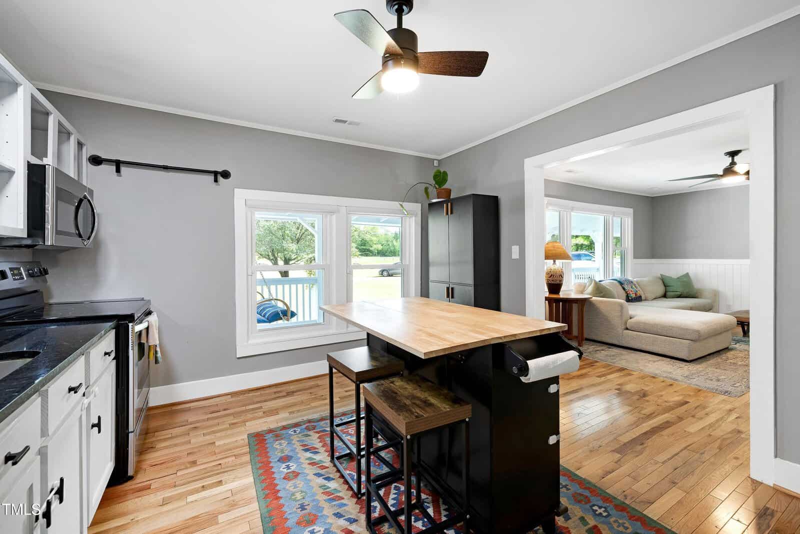 Modern kitchen with a wooden island, two stools, and stainless steel appliances, opening to a cozy living area with a sofa, rug, lamp, and large windows overlooking greenery.