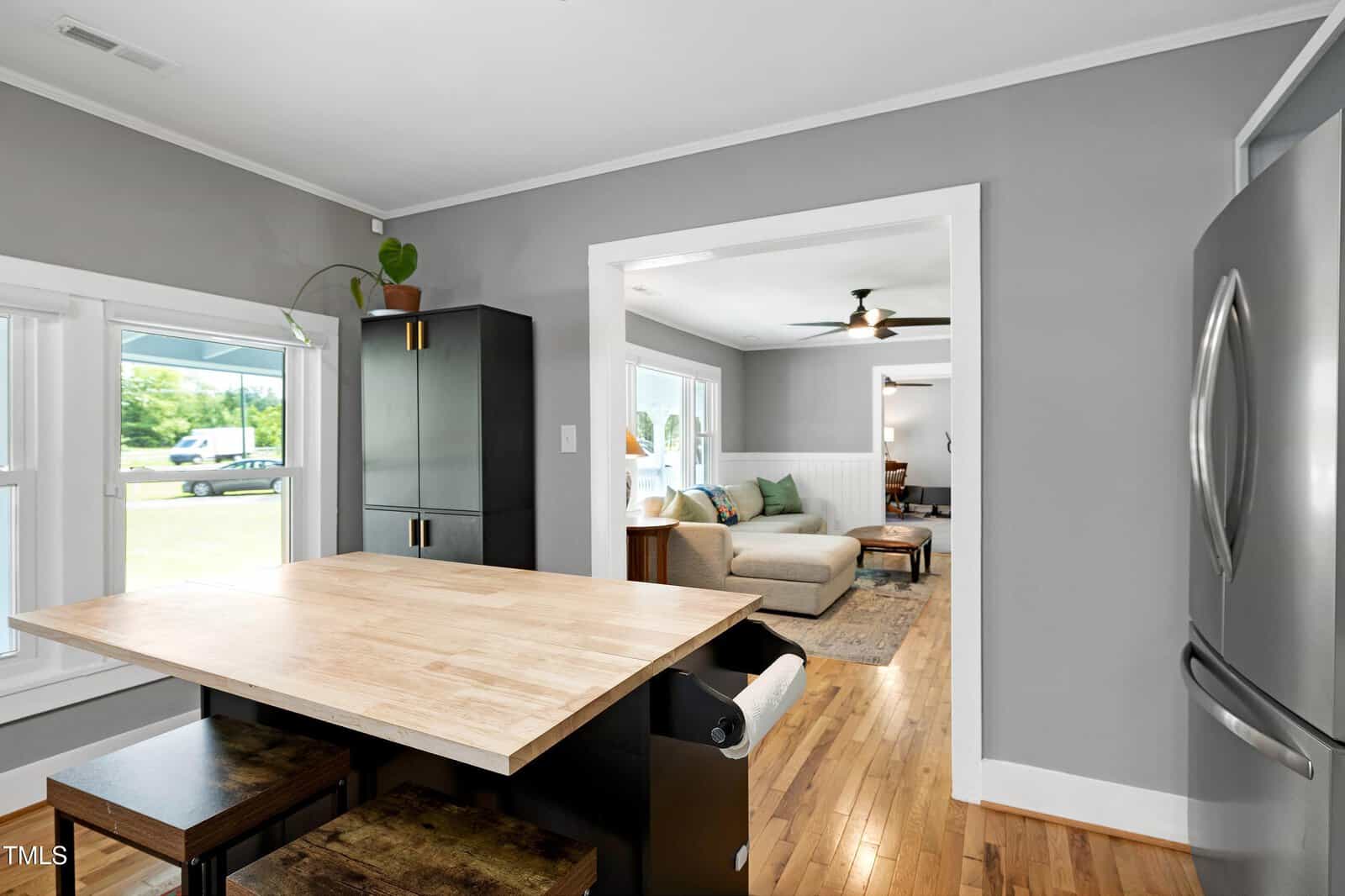 Modern kitchen with a wooden island, two stools, black cabinets, stainless steel fridge, and a view into a bright living room with a gray sectional sofa and a ceiling fan.