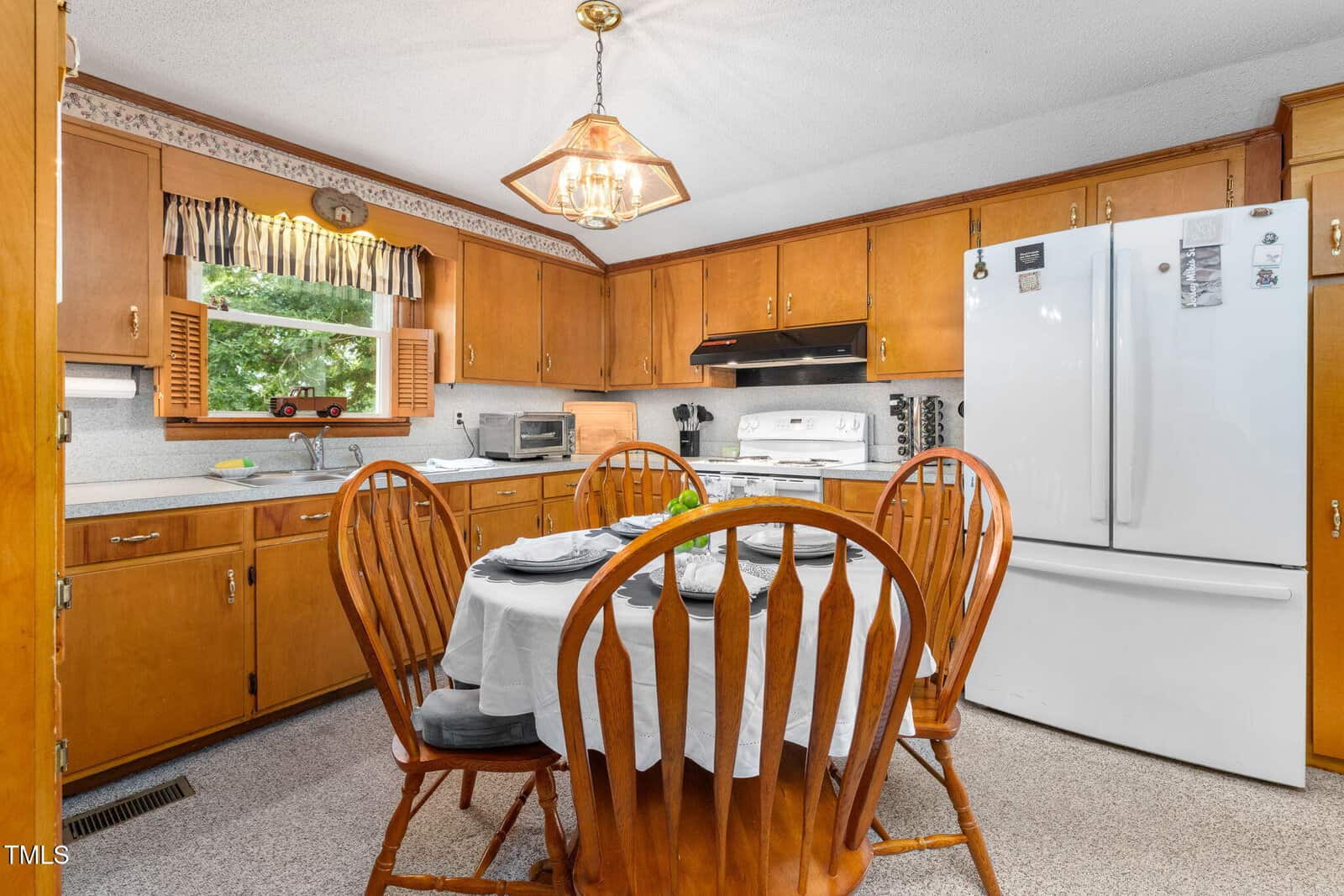 A cozy kitchen with wooden cabinets, a round table set for four, white appliances, and a window with a valance above the sink. Natural light brightens the space.
