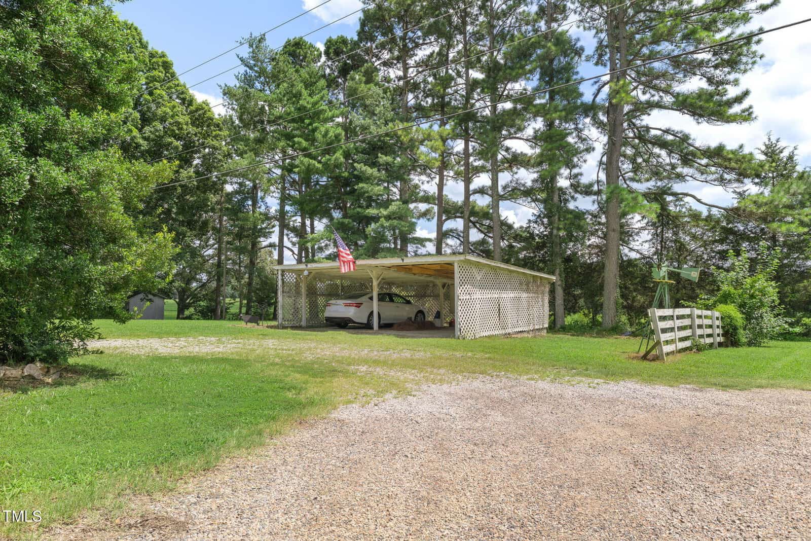 A white car sits under a large wooden carport with lattice sides. An American flag hangs from the roof. Tall pine trees and a grassy lawn surround the area under a partly cloudy sky.