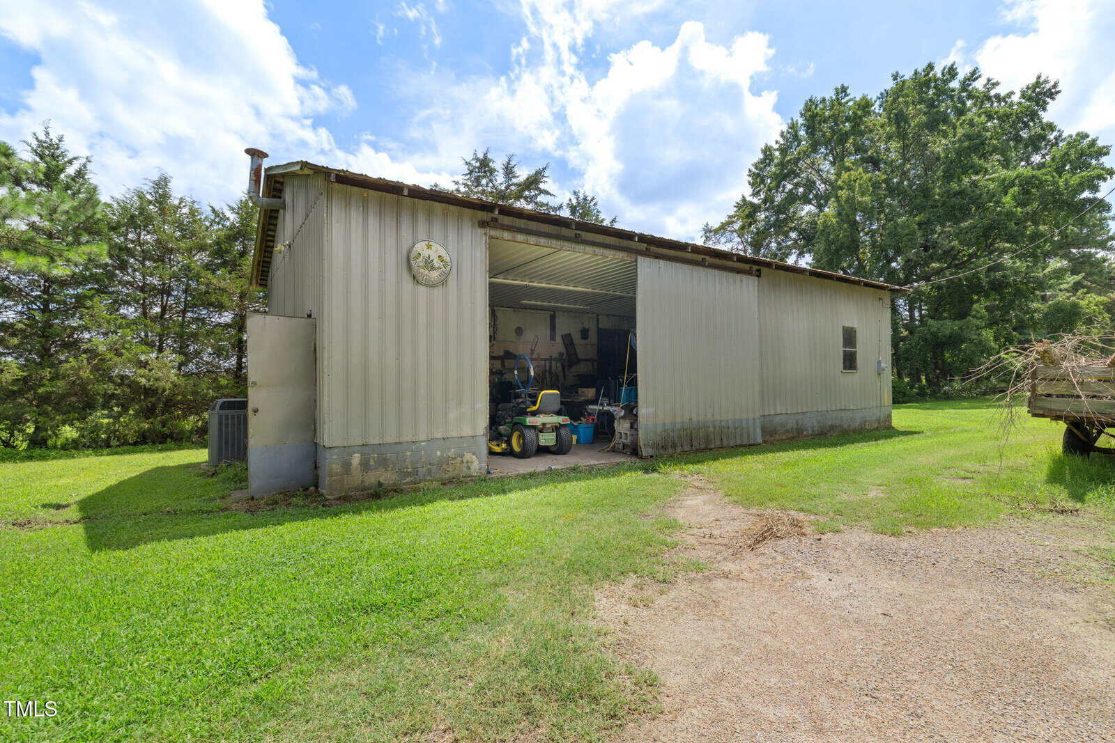 A metal shed with a partially open garage door reveals tools and equipment inside. The shed is surrounded by grass, trees, and a dirt driveway under a partly cloudy sky.