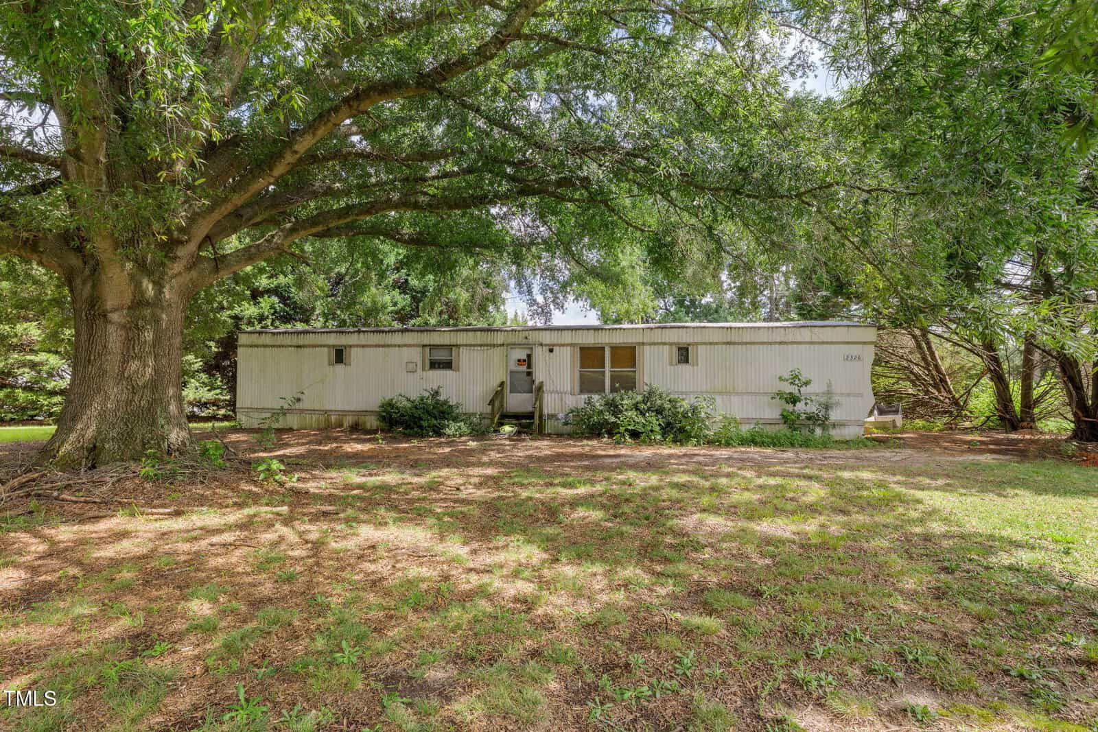 A white single-wide mobile home with a small front porch sits under a large, leafy tree, surrounded by grass and shrubs, on a sunny day.