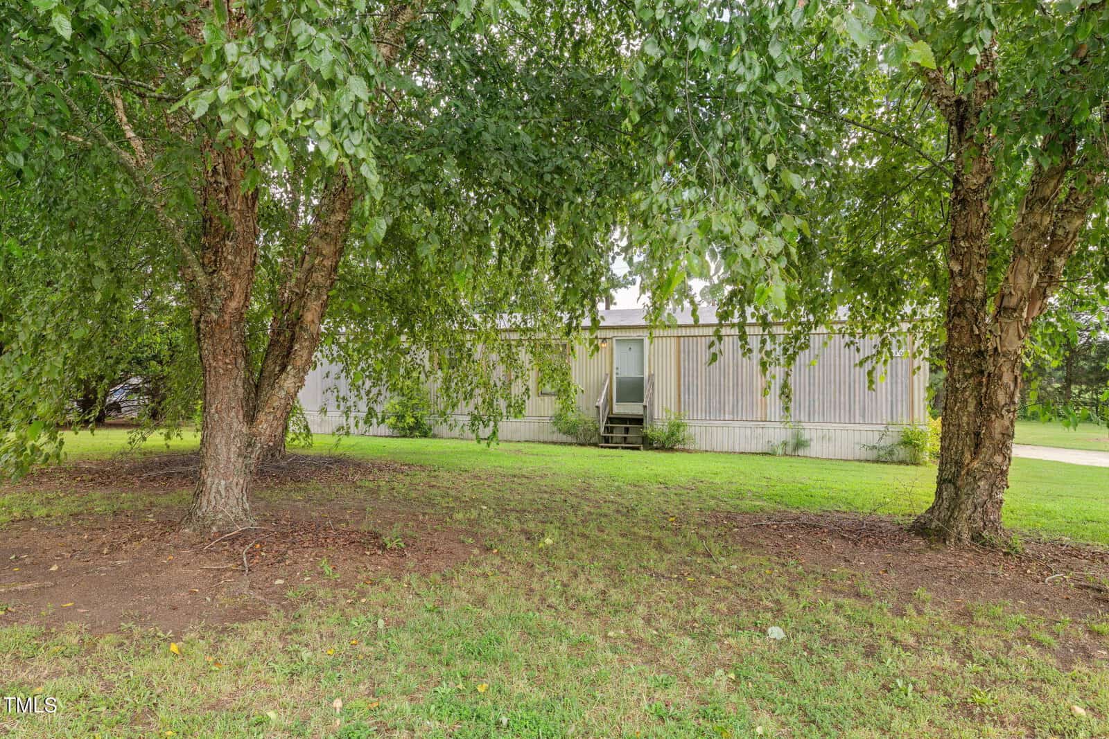 A single-wide mobile home with white siding is partially obscured by two large trees in the foreground. There are steps leading to the door, and the home sits on a grassy lawn with some patches of bare soil.