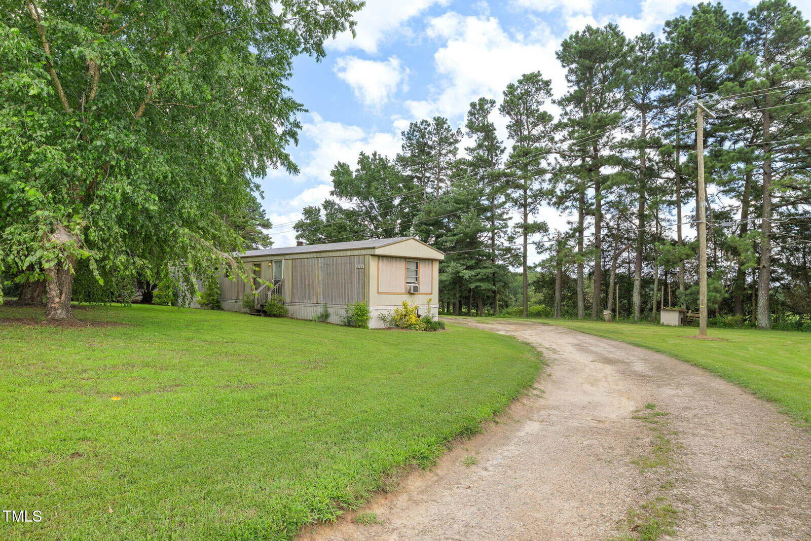 A single-wide mobile home sits on a grassy lot beside a curved dirt driveway, surrounded by trees under a partly cloudy sky.