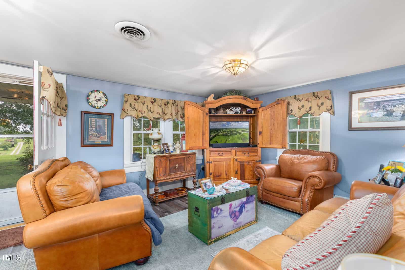 Cozy living room with tan leather chairs, a wooden cabinet holding a TV, a rustic side table, blue walls, and windows with patterned valances. Art and decor brighten the space. Door is slightly open to a grassy yard.