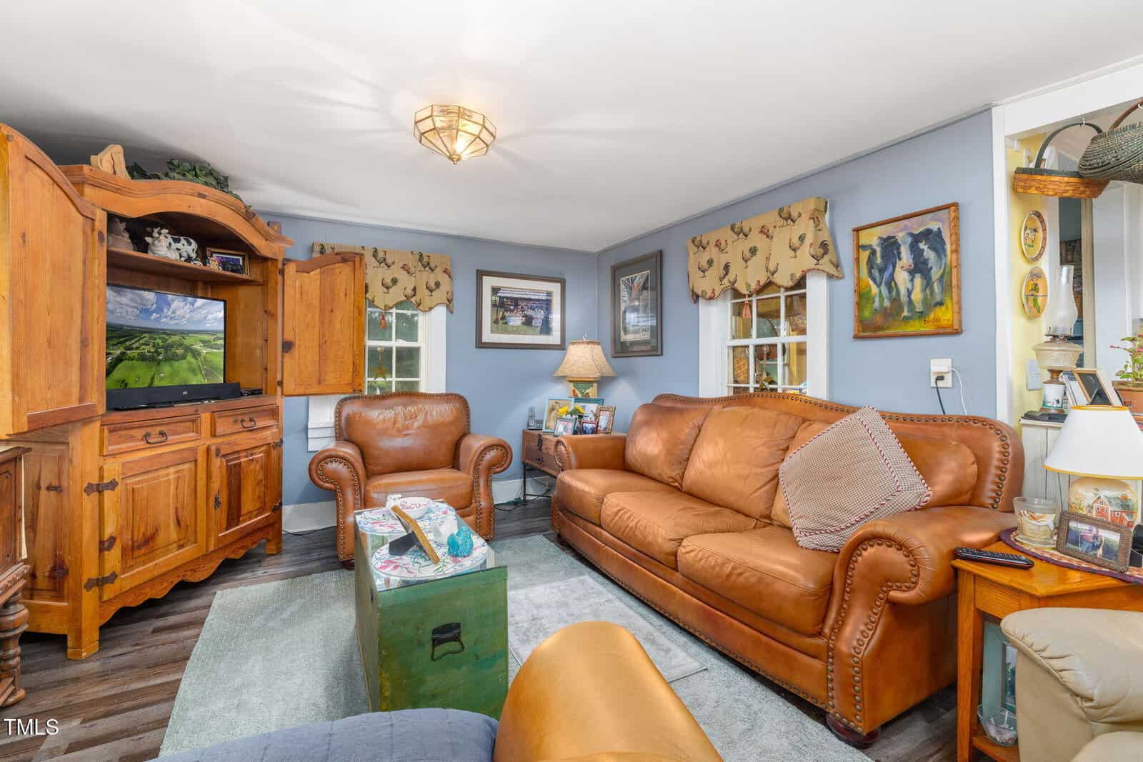 Cozy living room with leather sofa and armchair, wooden cabinet with TV, blue walls, art on walls, and windows with patterned valances. Coffee table and side table with lamp add warmth to the inviting space.