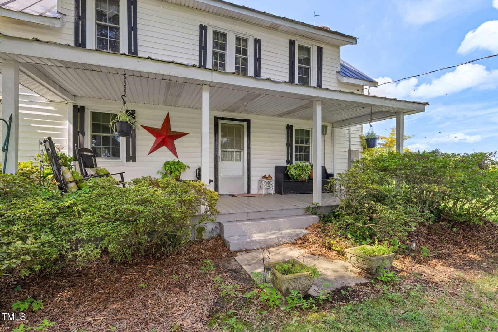 A white two-story house with black shutters and a covered front porch. The porch has plants, a bench, a rocking chair, and a large red star decor. Bushes line the walkway leading to the steps.