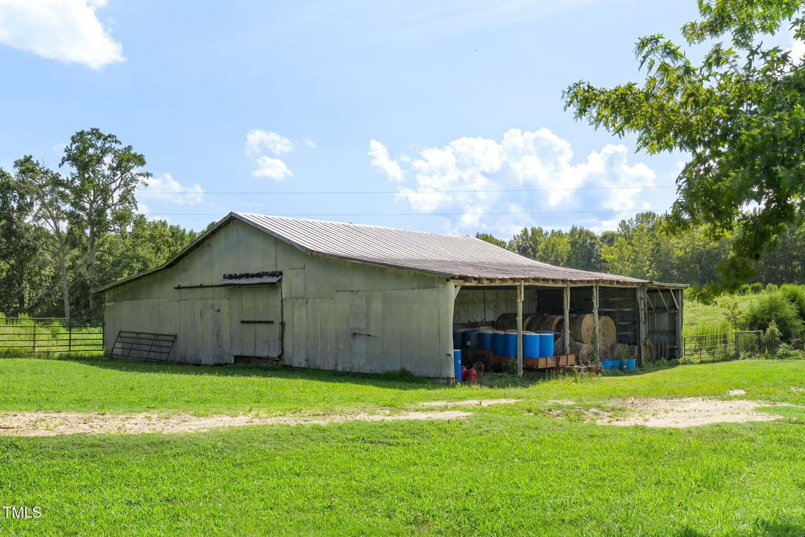 A weathered wooden barn with a metal roof stands on a grassy field. The barn has open sides with barrels, hay bales, and various equipment stored inside. Trees and blue sky with clouds are in the background.